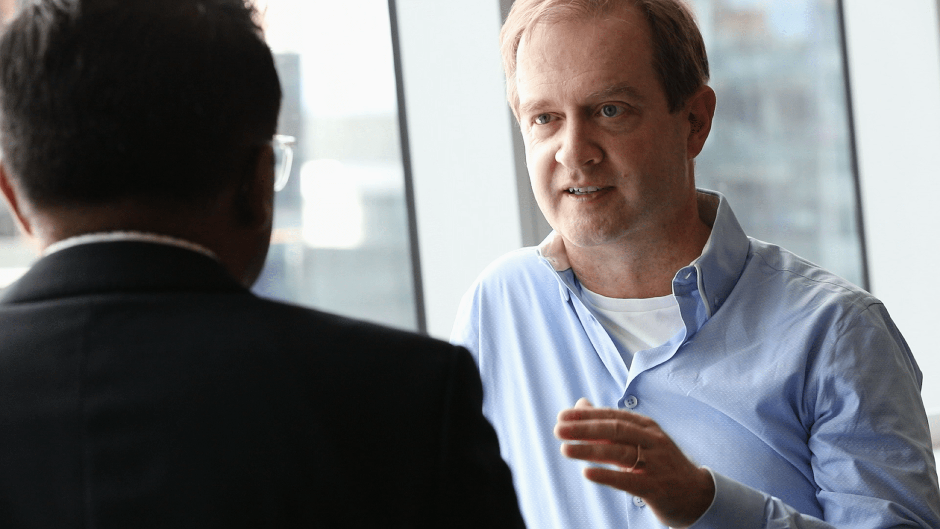 Adrian Kielhorn wears blue shirt and speaks to colleague in front of window